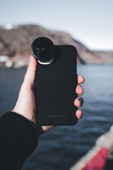 A close-up of a hand holding a smartphone with a fisheye lens attachment by a body of water.