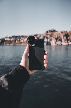 Person holding a smartphone with lens overlooking water and rocky landscape.