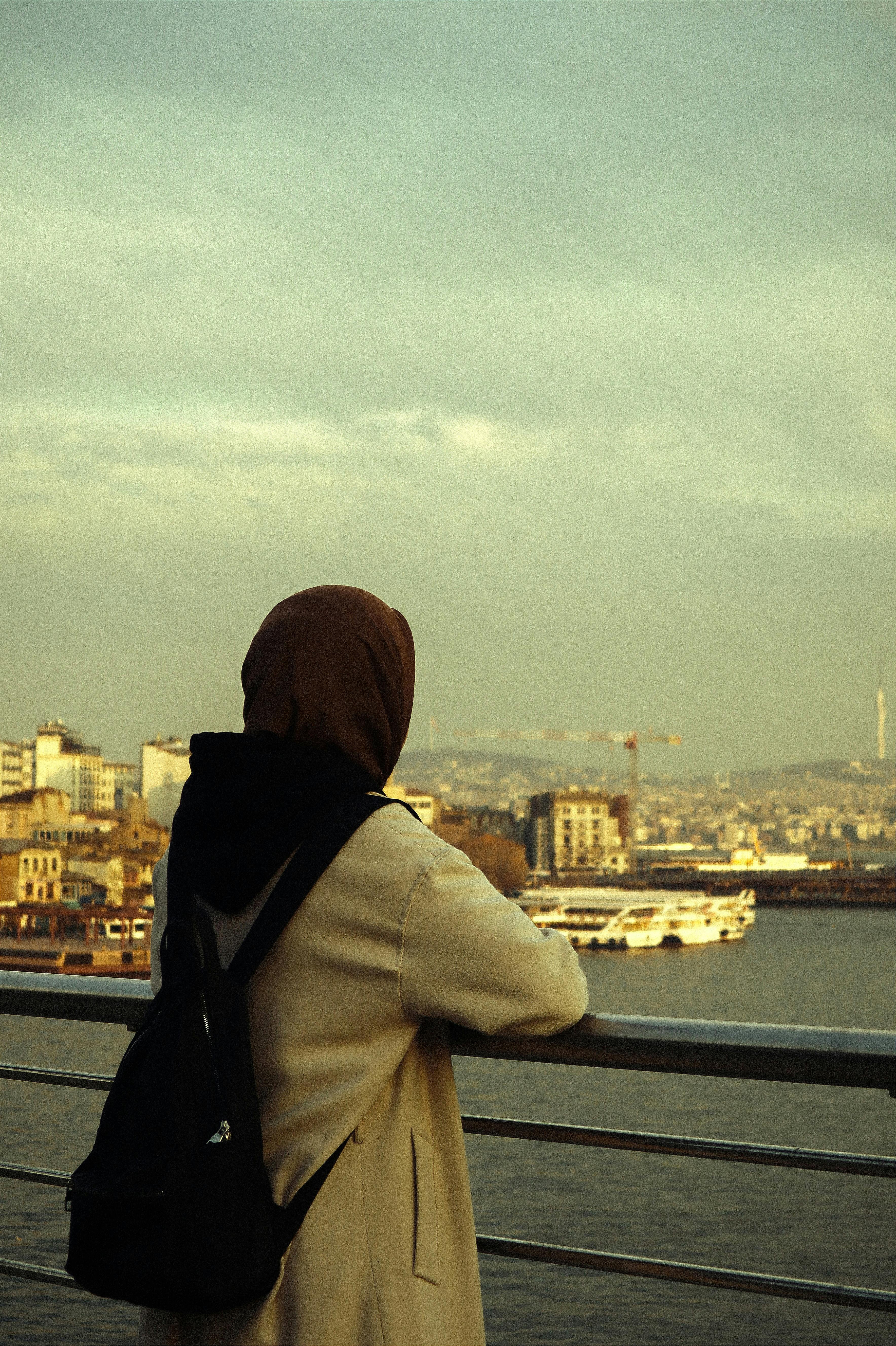 A woman in a hijab with a backpack stands on a railing, gazing at the cityscape by the water.