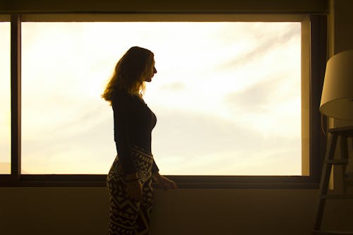 Женщина в черной рубашке с длинным рукавом и леггинсах в этническом стиле стоит у окна днем