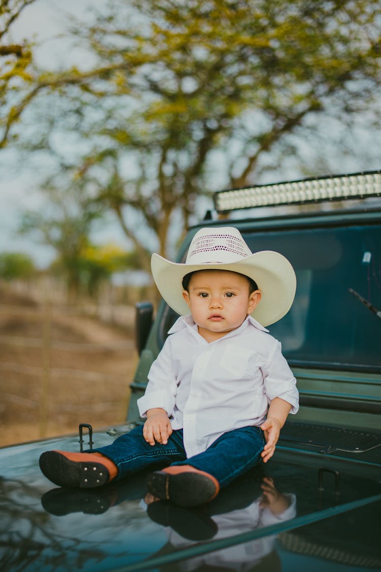 A Little Boy In A Cowboy Hat Sitting On A Car Hood 