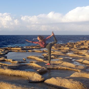 A woman performs a balance stretch on the rocky coast of Żebbuġ, Malta, during the day.
