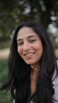 A portrait of a smiling woman with long black hair in an outdoor setting.