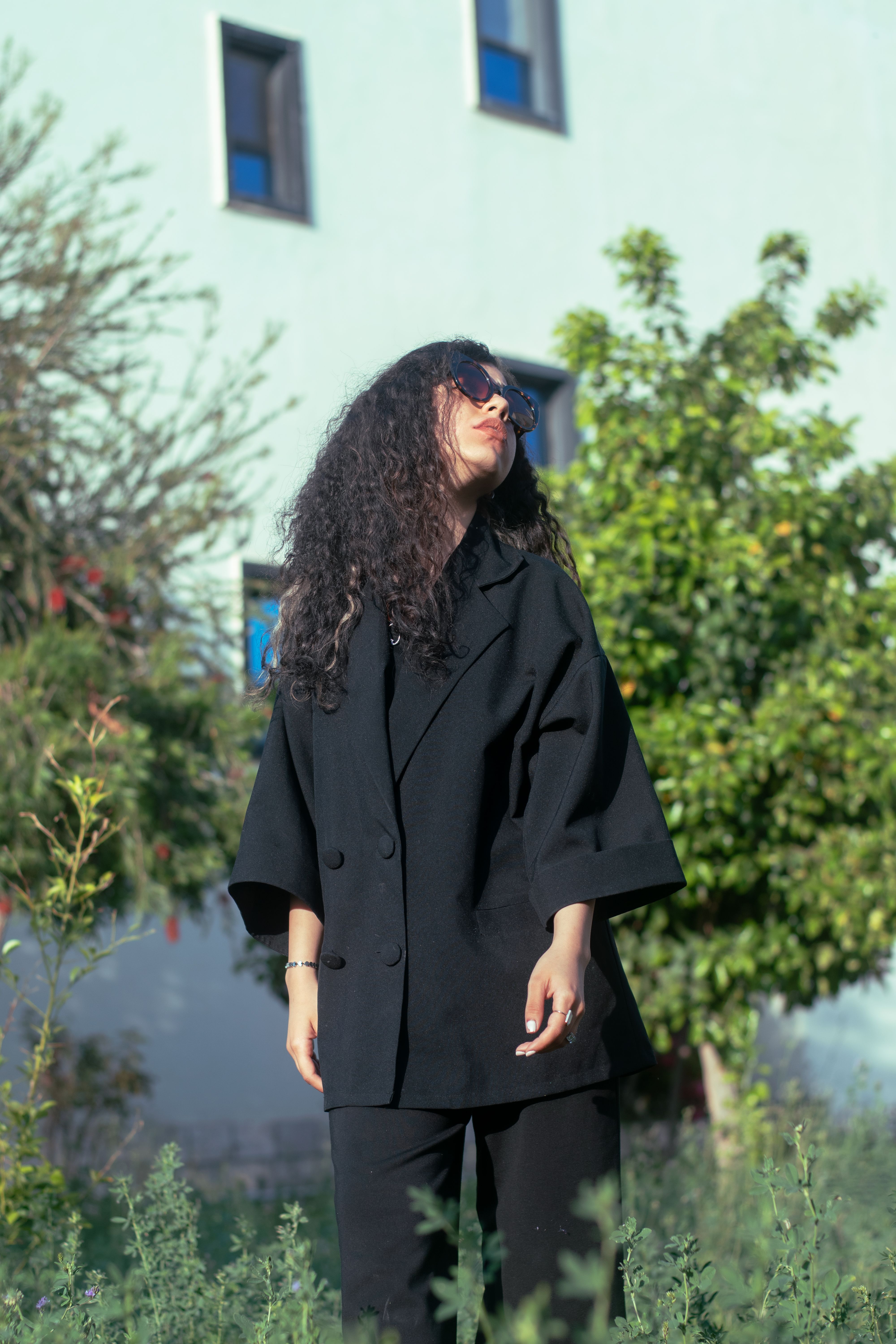 Fashionable young woman in a black blazer posing outdoors on a sunny day with green foliage.