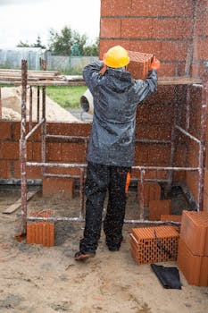 Worker with hard hat arranging bricks on a rainy construction site outdoors.