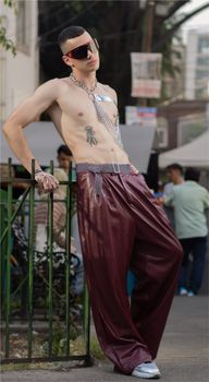Shirtless man in stylish sunglasses and pants poses outdoors in urban setting.