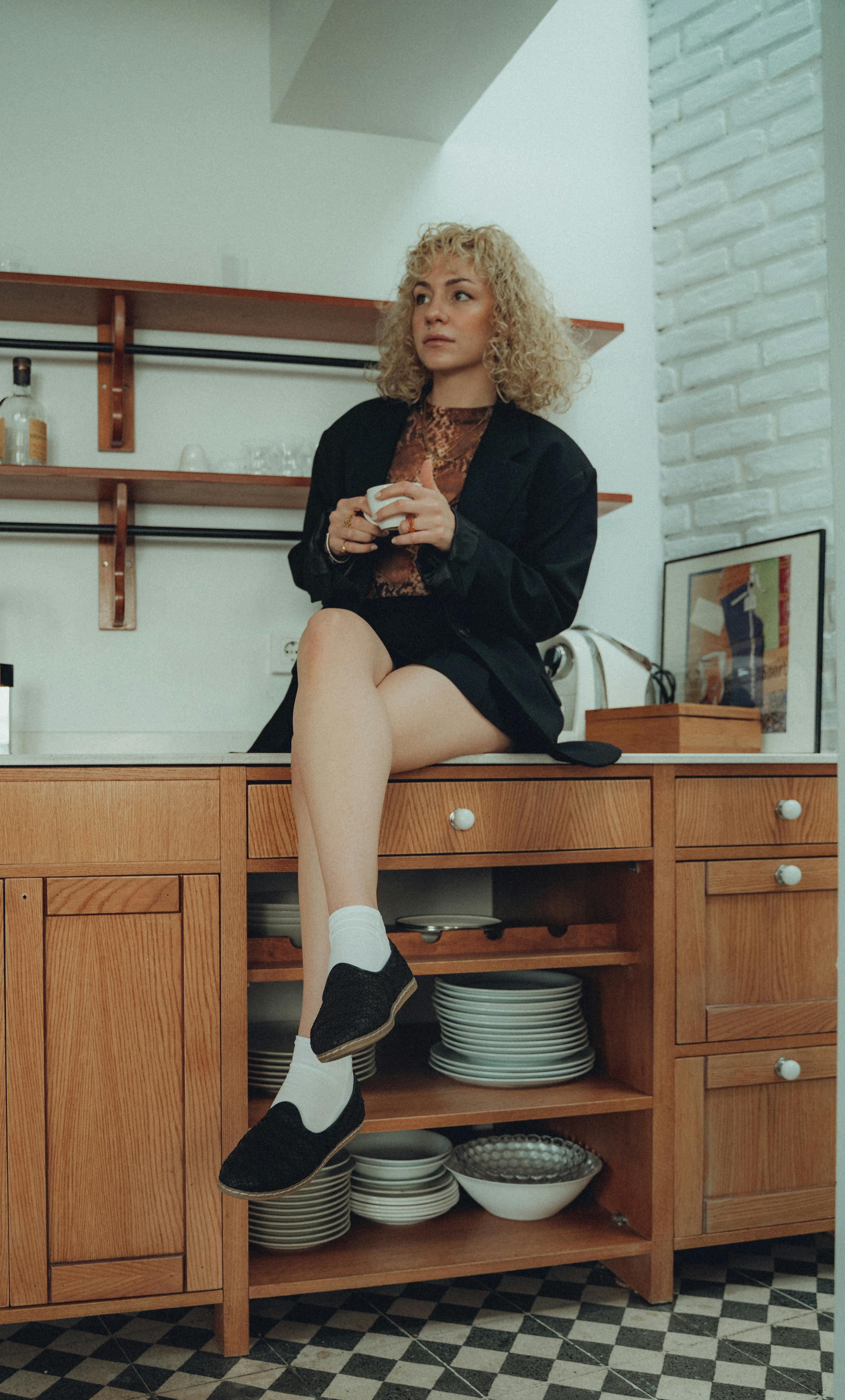 Elegant woman with curly hair posing in a stylish kitchen setting, holding a cup.