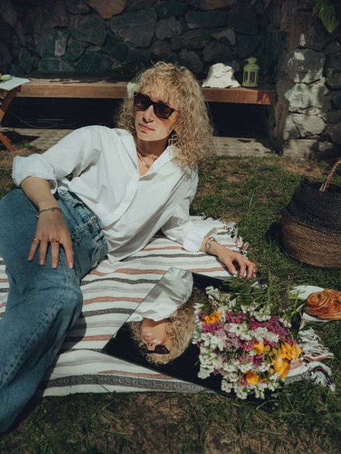 A woman in sunglasses relaxes outdoors on a striped blanket, surrounded by flowers and a mirror, enjoying the summer sun.