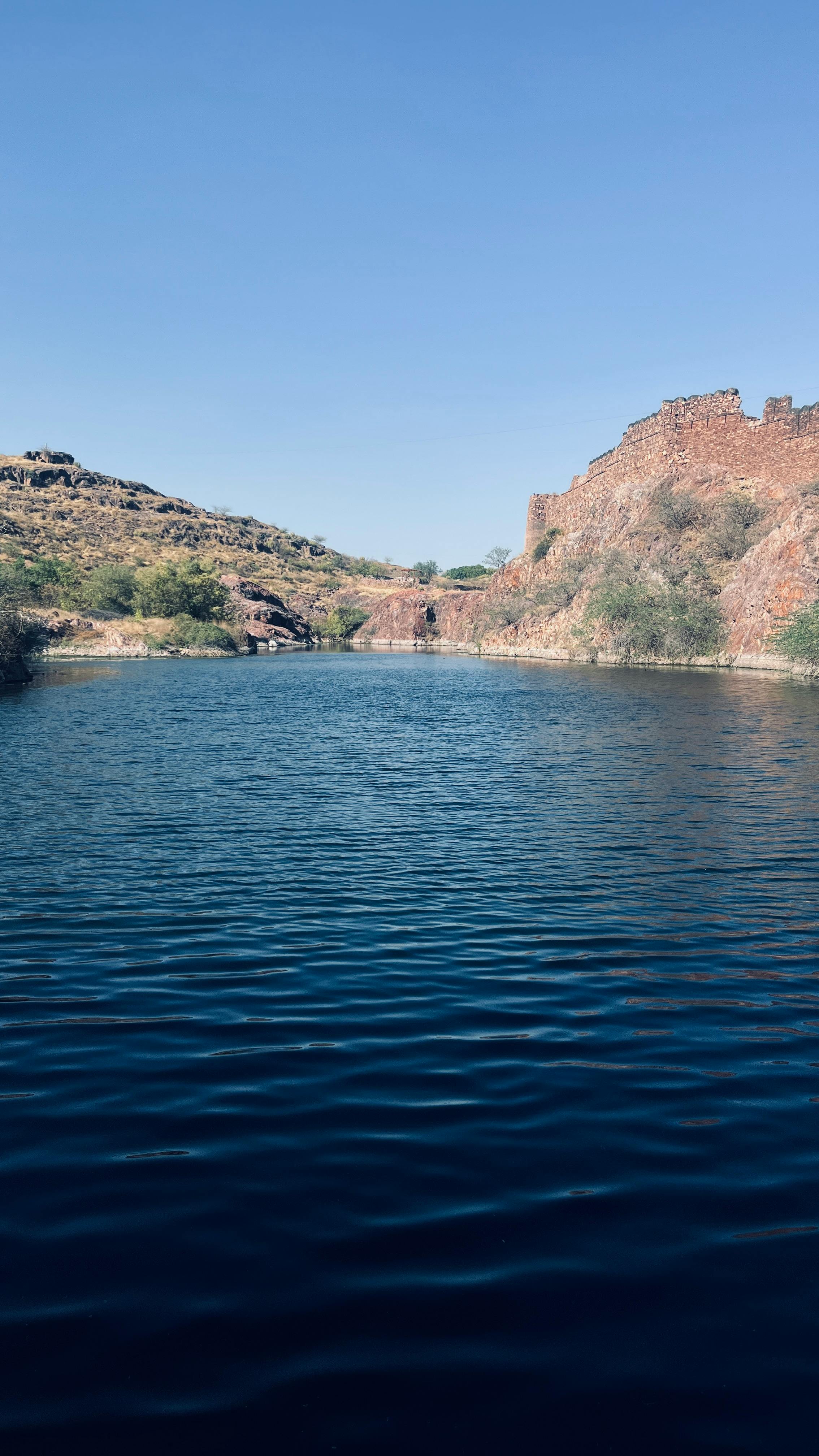 Tranquil lake surrounded by rocky hills under a clear blue sky in Jodhpur, India.