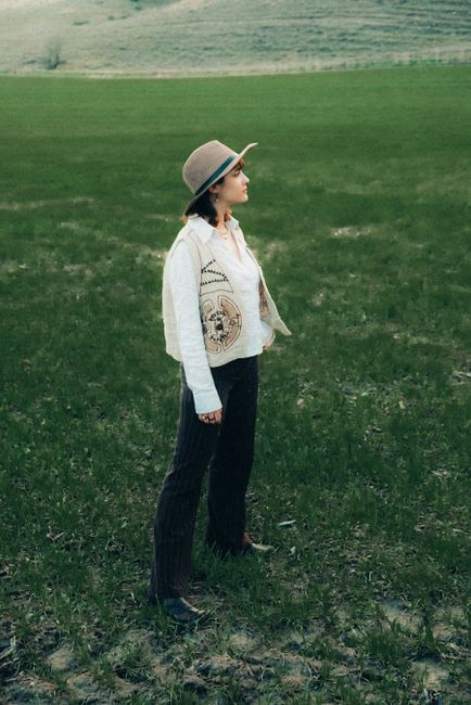 A young woman in a hat and vest poses in a lush green field, exuding fashion and style.