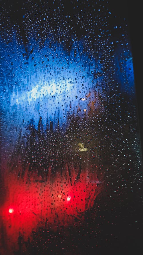 Close-up of a Window with Raindrops and Blue and Red Lights behind the Glass