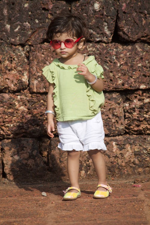 A Little Girl in a Summer Outfit and Red Glasses Standing by a Wall 