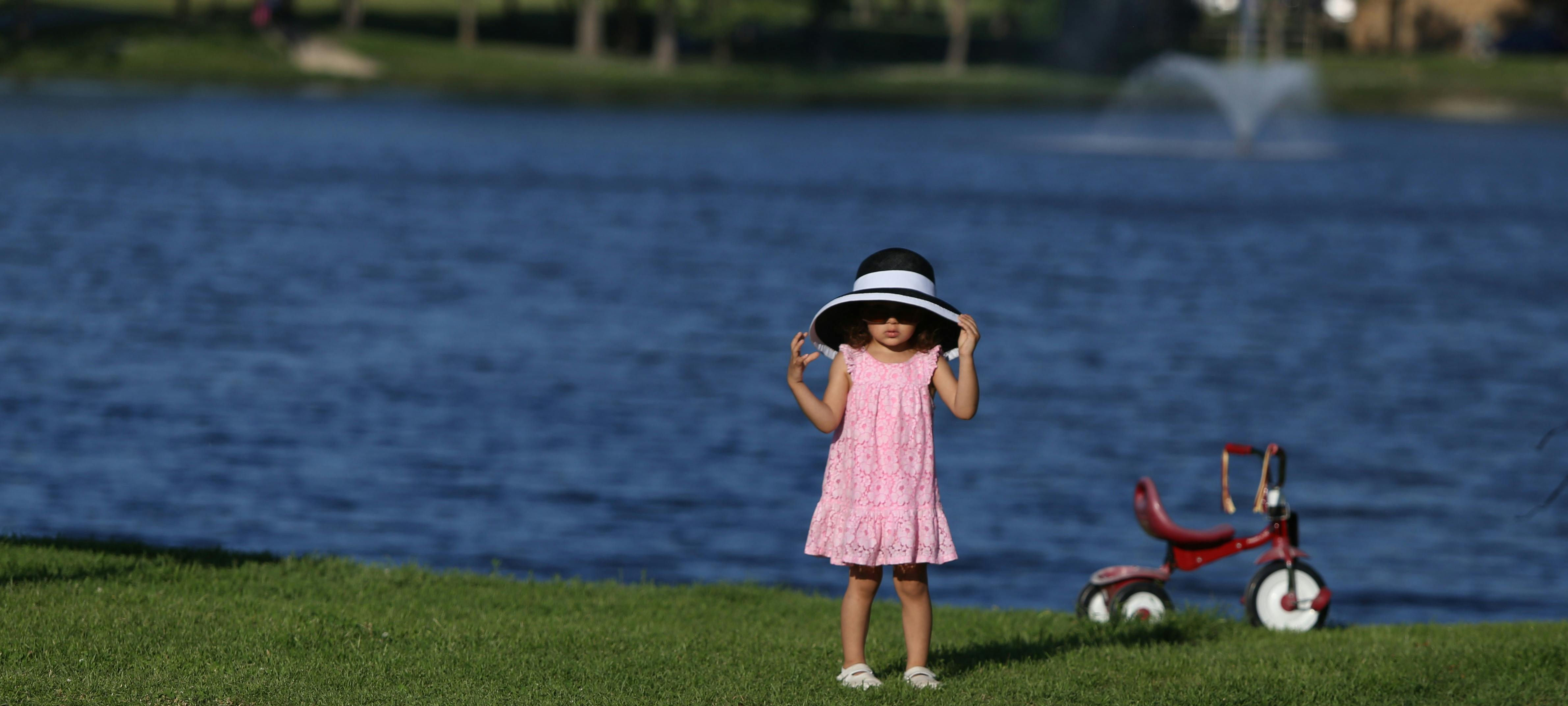 Kid in Pink Dress With White and Black Sun Bucket Hat Beside the Red Pedal Trike in Front of Body of Water