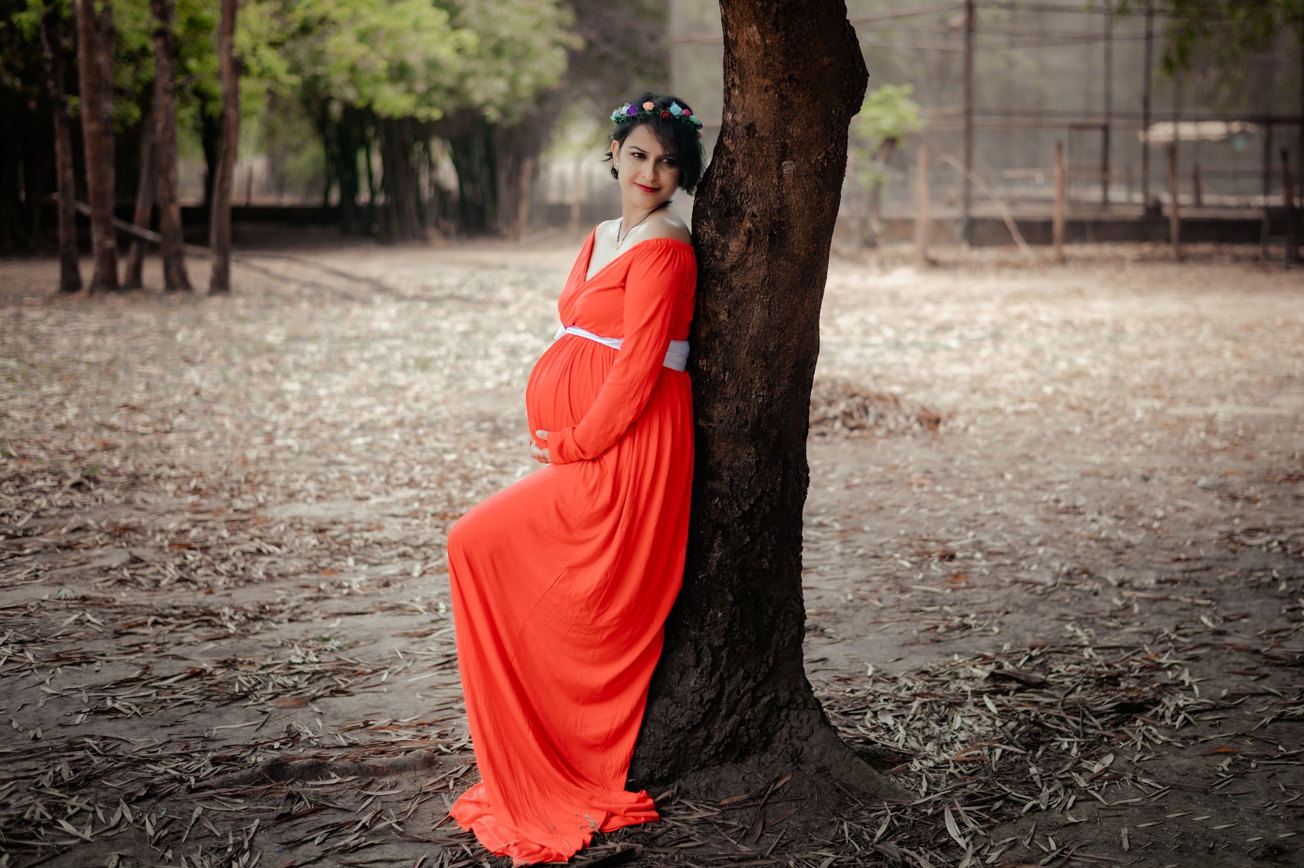 Pregnant woman in red dress leans against a tree in the park, exuding elegance and warmth.