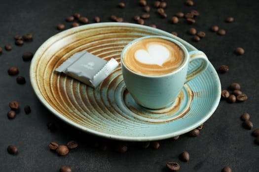 A steaming cup of latte with heart-shaped froth art on a ceramic plate with coffee beans scattered around.
