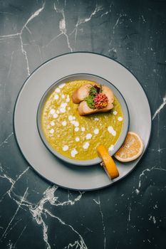 Stylish flat lay of a gourmet pea soup topped with crostini and a lemon slice on a marble table.