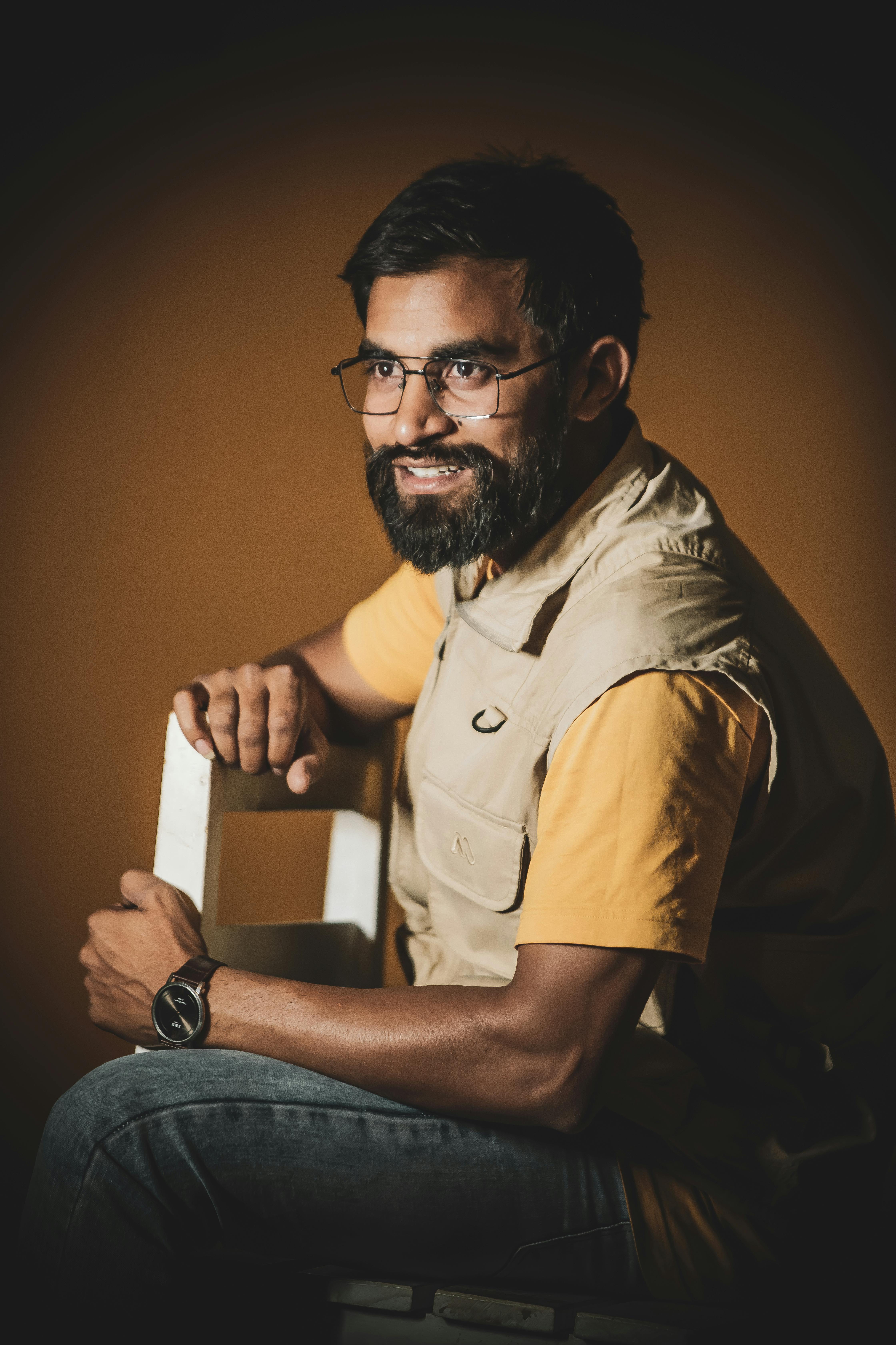 Portrait of a mid adult man with beard and glasses, sitting against brown background.