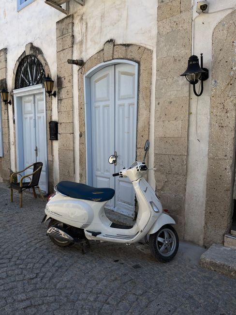 White motor scooter parked by vintage doors on cobblestone street in Alaçatı, İzmir, Türkiye.