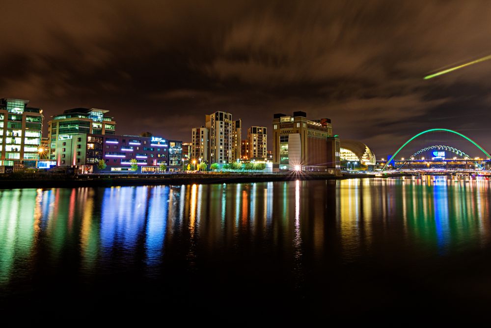 Vibrant city skyline at night reflecting on water with colorful lights and a modern bridge.