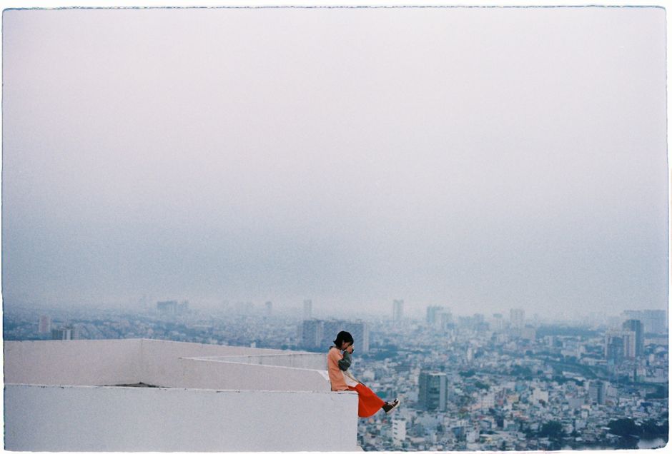 A woman sits poised on a building's edge, high above a sprawling cityscape.