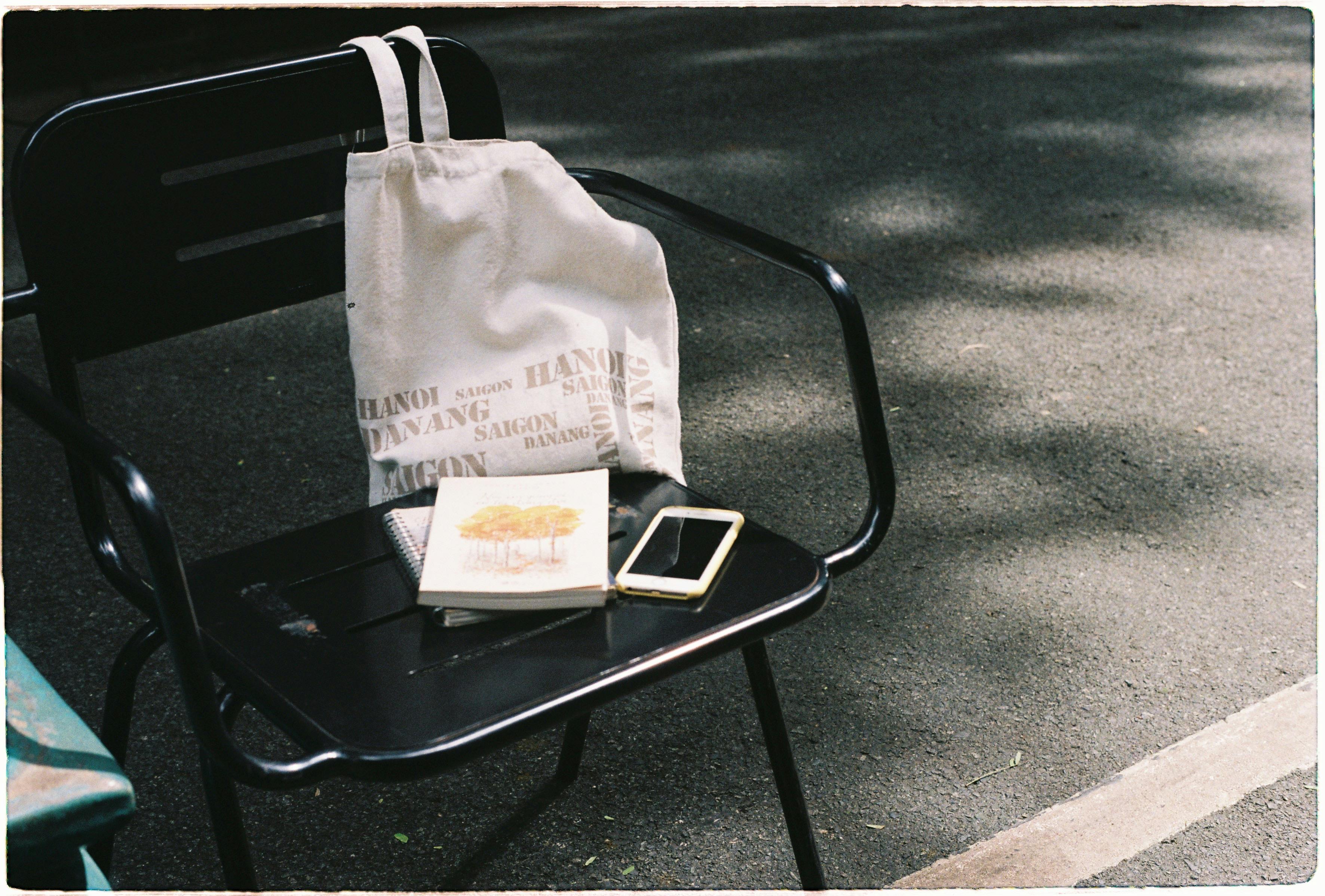 A casual street scene showing a chair with a bag, notebook, and cellphone on a sunny day.