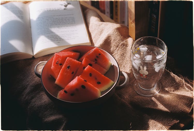 A Bowl With Watermelon, A Book And A Glass Of Water On A Table 