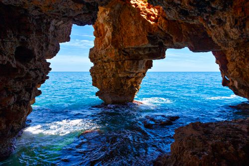 Free Rocky cave opening to the vibrant Mediterranean Sea on a sunny day. Stock Photo