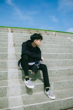 Young man sitting on steps outdoors in casual fashion wear under blue sky.