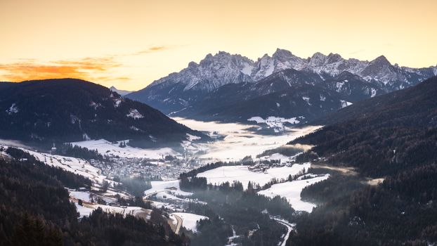 Breathtaking view of the snow-covered Dolomites at sunrise with fog and village below.