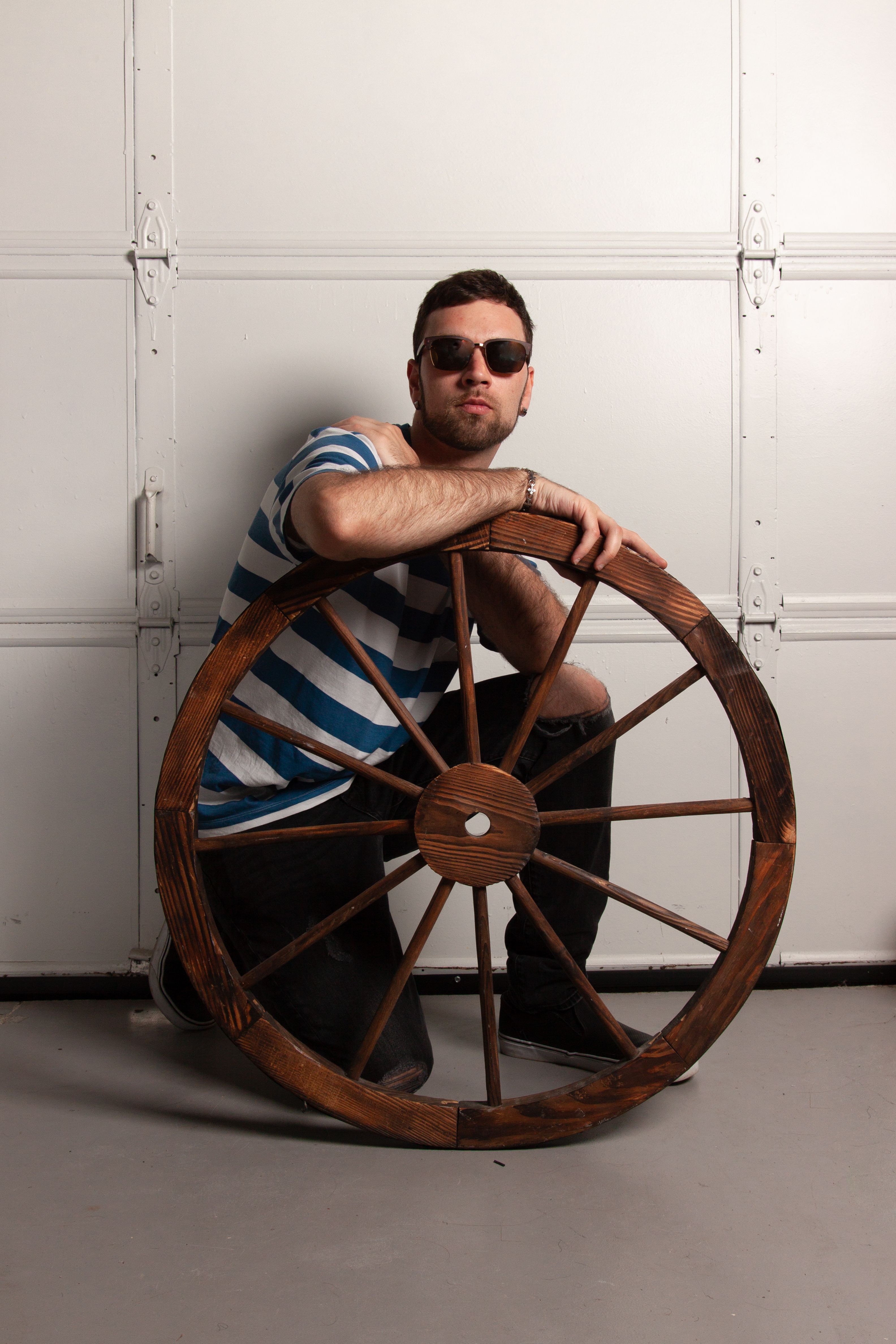 A man with sunglasses kneeling and posing with a vintage wooden wheel indoors against a white wall.