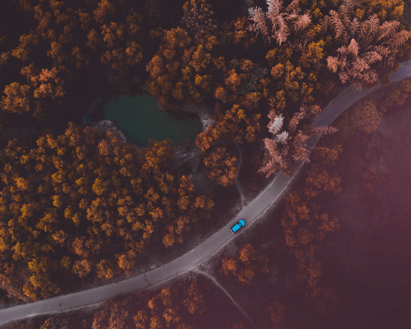 Drone shot capturing a vibrant blue car on a road amidst lush autumn trees.