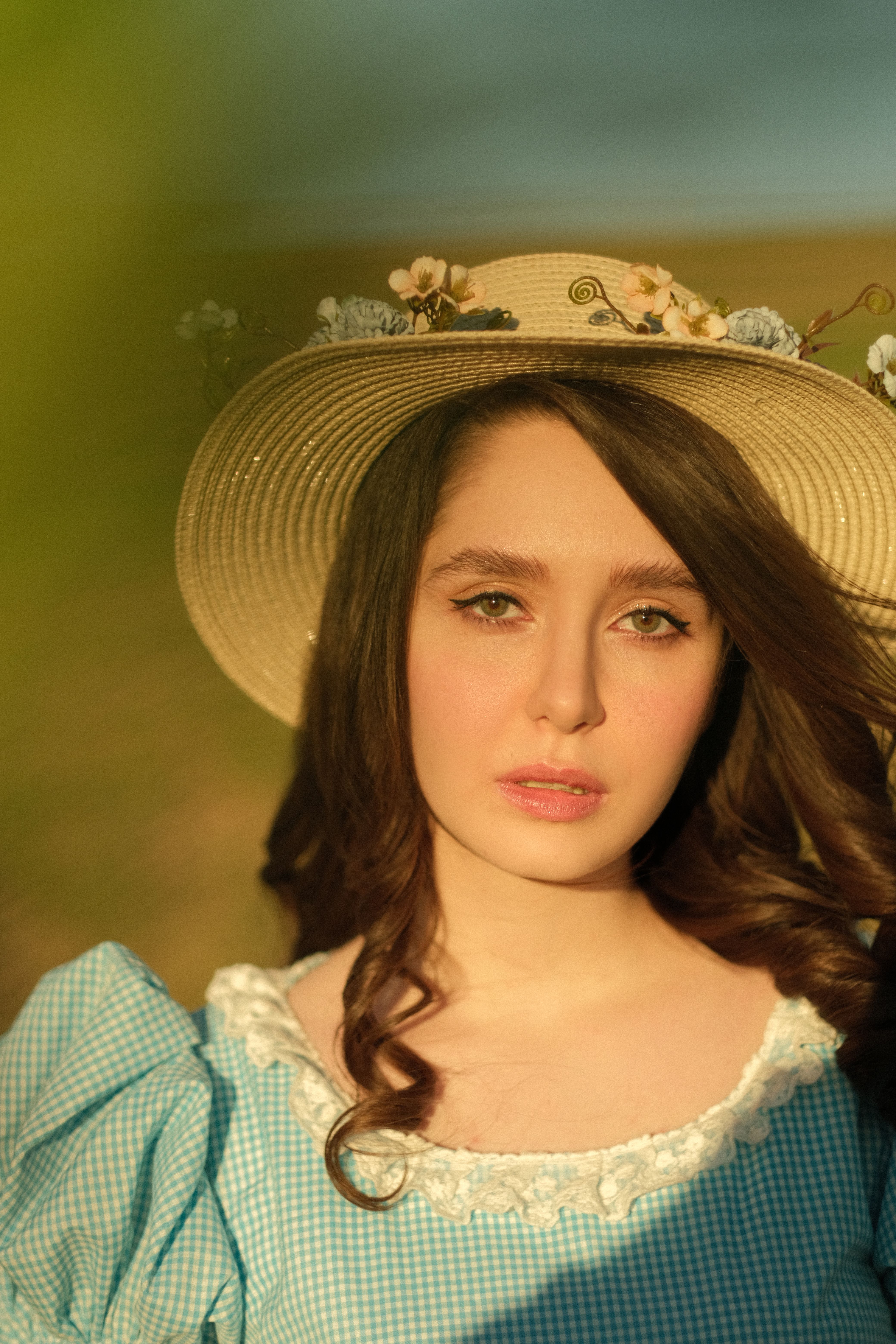 Portrait of a woman in a floral hat and blue dress, sunlit in a rural setting.