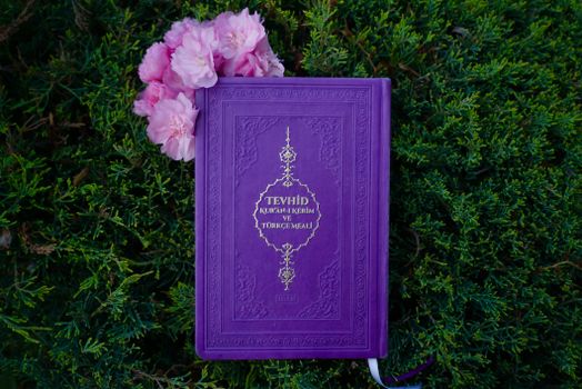 A purple Quran rests on vibrant green shrubs, adorned with pink flowers.
