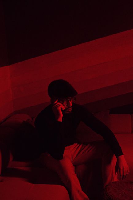 A man in a room with red ambient lighting, sitting on a couch, seemingly in contemplation.