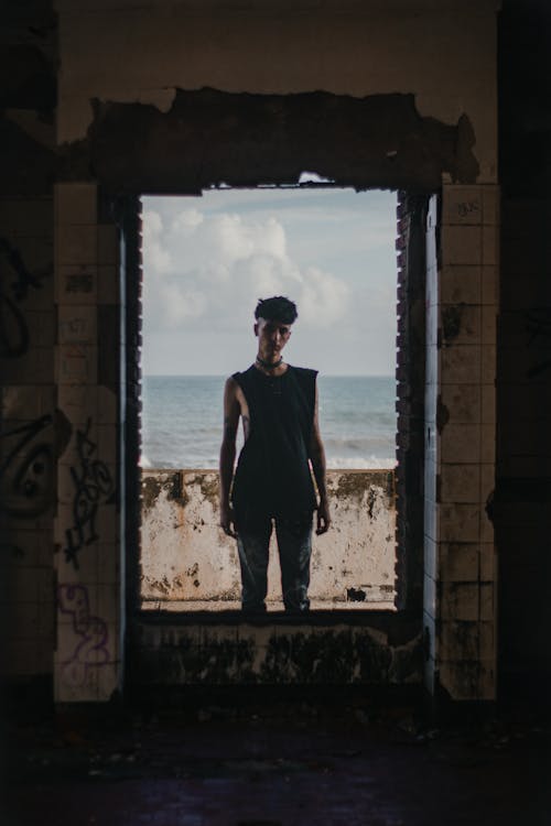 Young Man in Black Tank Top Standing in Window of Abandoned Building