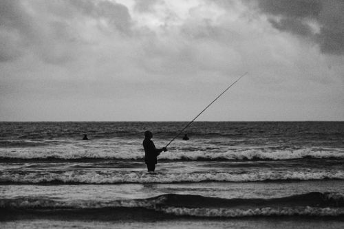Free Silhouette of a man fishing at the shore under an overcast sky, with waves crashing around. Stock Photo