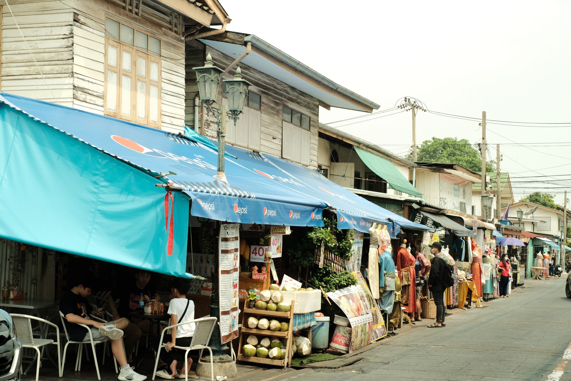 Lively city market street with various stalls and shoppers under bright blue awnings.