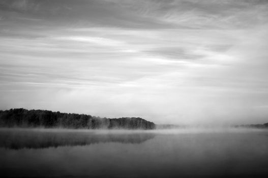 A serene black and white image of misty forest reflecting on a tranquil lake.