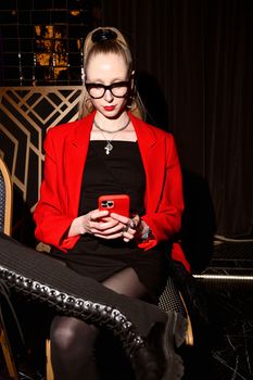 Chic woman in a red jacket and black dress using smartphone indoors. Stylish modern fashion.