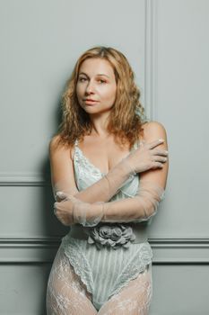 A stylish woman poses confidently in elegant lingerie against a light gray wall.