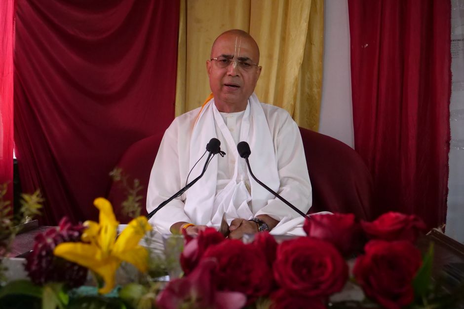 A religious leader in white speaks surrounded by vibrant flowers indoors.
