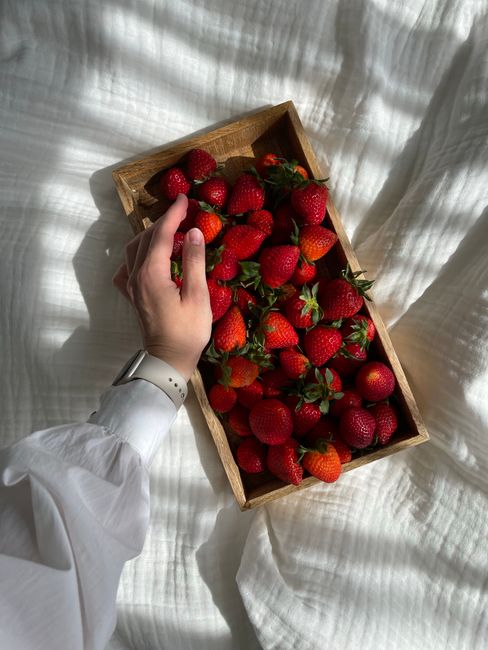 Hand reaching for fresh strawberries in a wooden box on a white fabric background, bright daylight.
