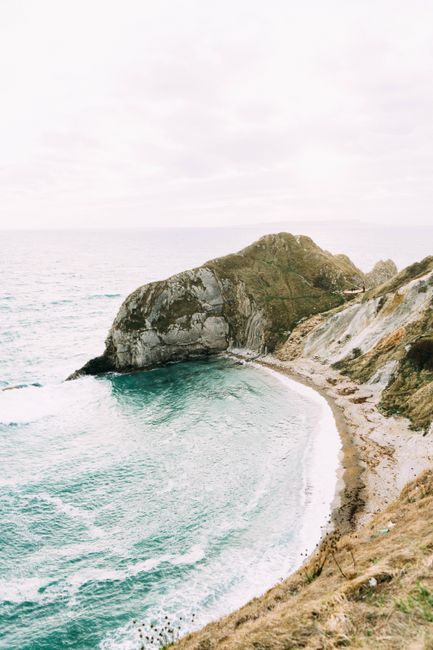 A scenic view of a coastal cliff and cove with turquoise waves crashing on the shore, captured from above.