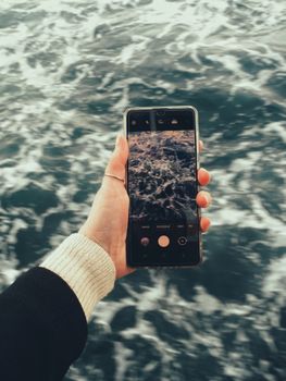 A close-up of a hand holding a smartphone recording ocean waves.
