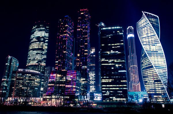 Vista noturna iluminada dos modernos arranha-céus de Moscovo, destacando as luminosas luzes da cidade e a arquitetura urbana.