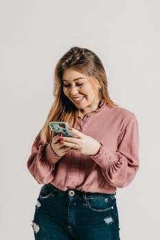 Young woman smiling while checking her smartphone, dressed casually in a pink blouse and jeans.