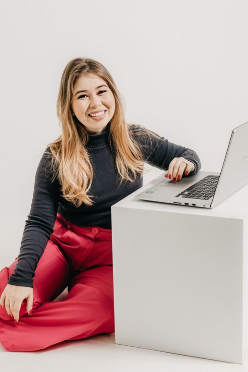 Free Young woman sitting next to a laptop in a studio, wearing red trousers. Stock Photo