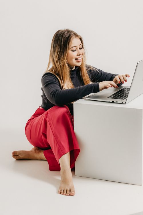 Free Portrait of a young woman in red trousers working on a laptop in a bright studio setting. Stock Photo