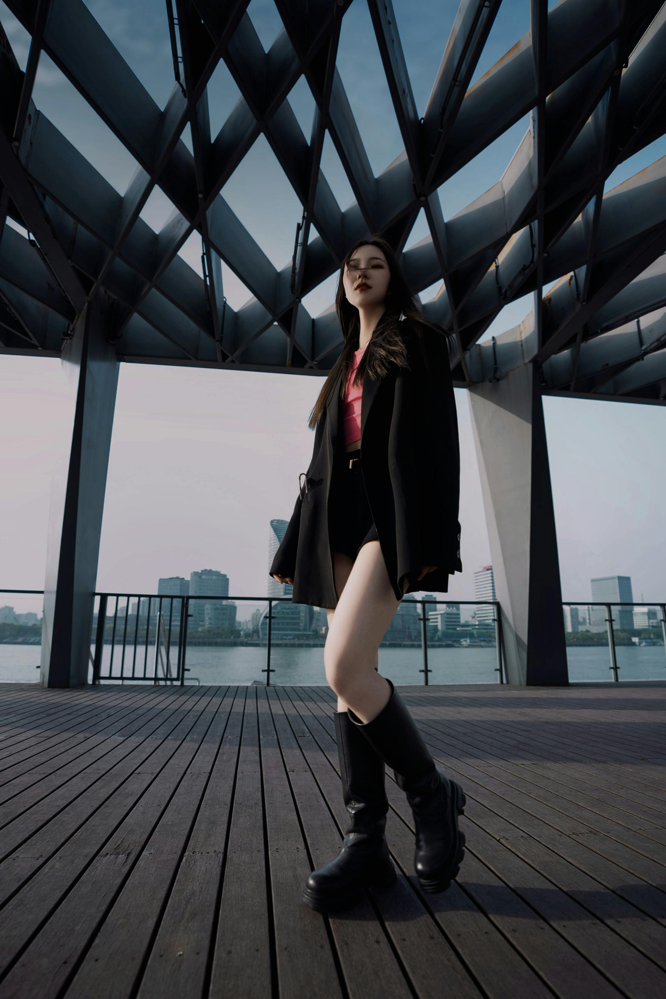 Stylish woman in a blazer and boots posing under modern urban architecture, showcasing contemporary fashion.