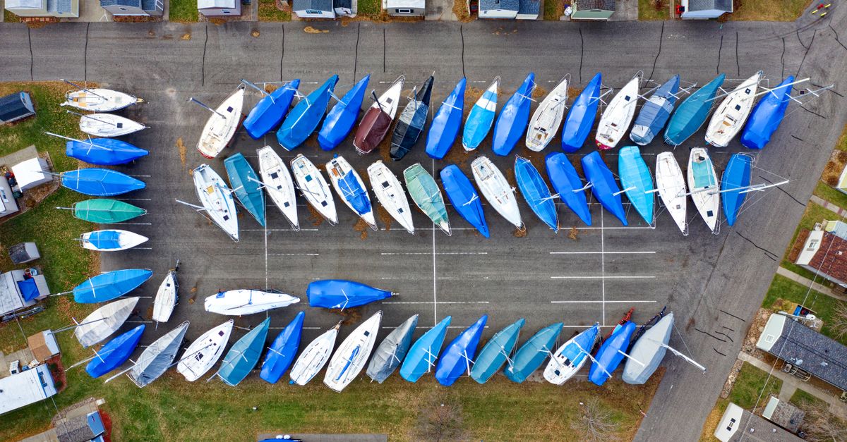 Aerial shot of numerous sailboats covered with blue tarps in an urban marina parking area.